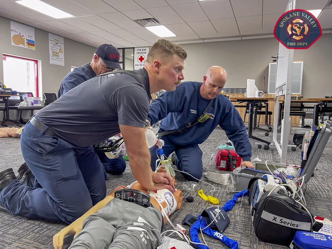 Emergency responders are performing CPR on a mannequin during a training session. The room includes medical equipment and a computer. A red emblem on the wall reads "Spokane Valley Fire Dept."