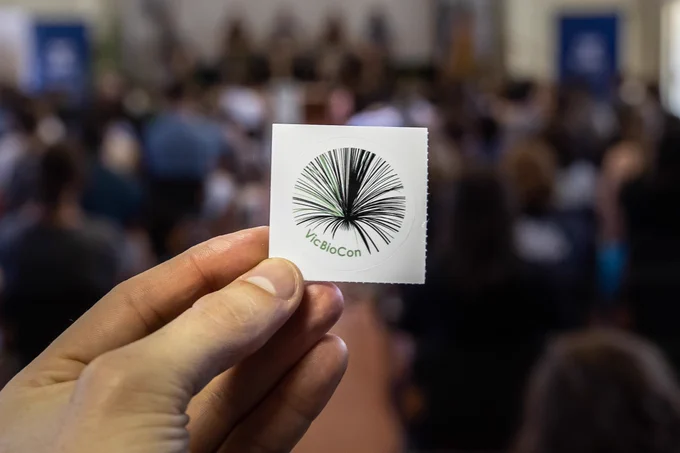 A photo of a hand holding up a sticker of the VicBioCon logo, with a blurry conference hall in the background