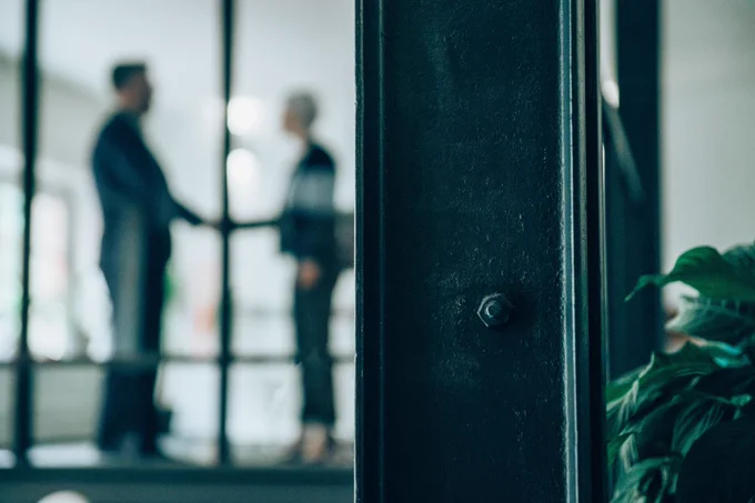 Two individuals having a conversation in a modern office space, partially obscured by a foreground black door frame.