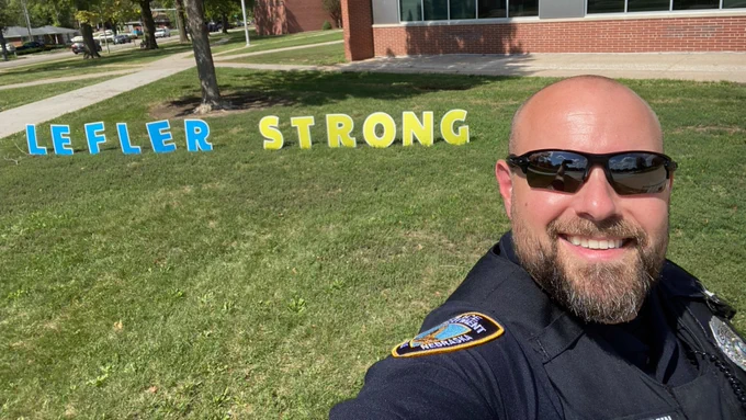 A person in a police uniform takes a selfie on a grassy area in front of a building. Large, colorful letters on the grass spell "LEFLER STRONG."