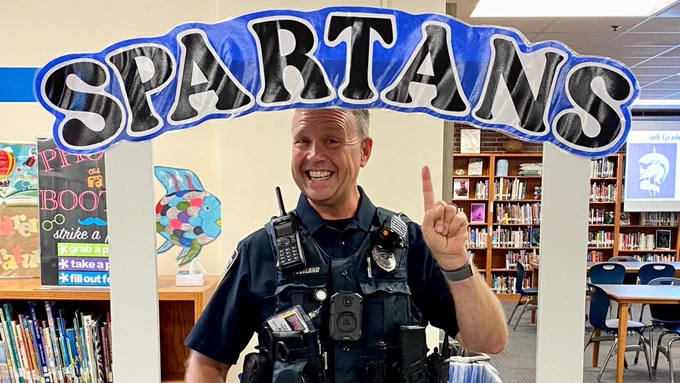 A person dressed in a police uniform stands inside a library. They are smiling and posing with one finger pointed upward through a decorative frame with the word "SPARTANS" on it. Bookshelves and various library decorations are visible in the background.