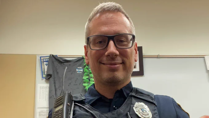A person wearing glasses and a police uniform with a badge and a radio, inside an office setting. A framed picture and a t-shirt are visible in the background.