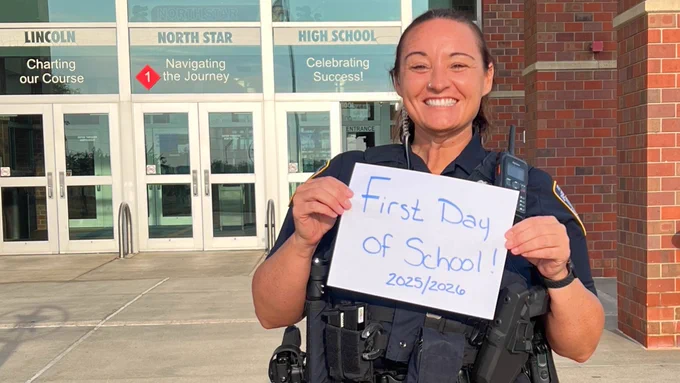 A person in a police uniform stands smiling in front of the entrance to Lincoln High School. They hold a sign reading "First Day of School! 2025/2026." The school's doors feature phrases like "Charting our Course," "Navigating the Journey," and "Celebrating Success."