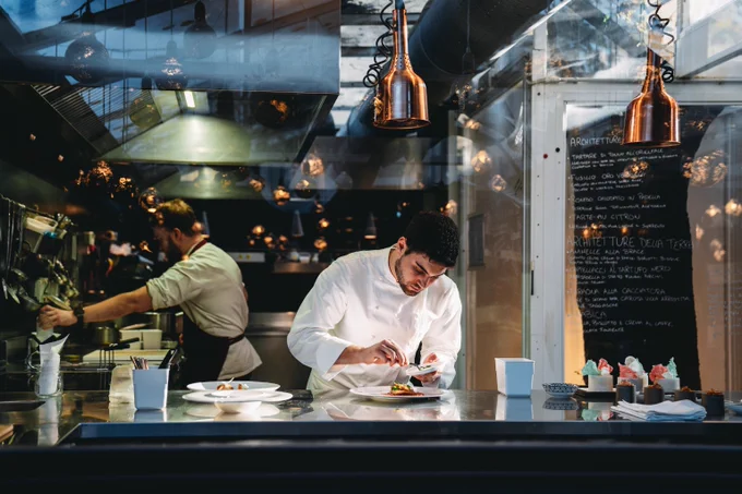 Chef carefully plating a gourmet dish in a modern restaurant kitchen with copper pendant lights overhead.