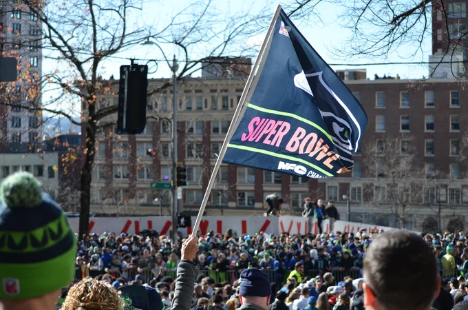 A large crowd is gathered outdoors with someone holding up a Seattle Seahawks flag that reads "Super Bowl" and "NFC Champion." People are dressed in Seahawks attire, and buildings are visible in the background.