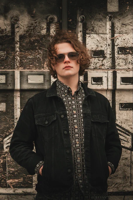 A professional vertical urban portrait of a young man with wavy, ginger-brown hair standing against a textured background. He wears silver aviator sunglasses and has a calm, confident expression. His outfit consists of a dark denim jacket worn open over a button-down shirt featuring a bold, repeating black and white square geometric pattern. The background is composed of weathered plywood with black painted markings and metallic rectangular accents, creating an industrial, street-style aesthetic. The lighting is soft and overcast, which minimizes harsh shadows and allows the intricate patterns of the shirt and the rough splinters of the wood to remain sharp and detailed. The composition is a medium shot, focusing on the subject from the waist up with his hands tucked into his jacket pockets, emphasizing a cool, relaxed posture that complements the gritty, modern environment.