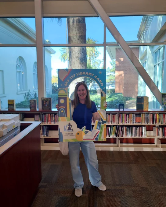 Person standing inside a library holding a colorful frame that says 'My 1st Library Card' with bookshelves and large windows in the background.