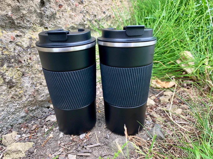 Two black travel mugs standing on some stonework in the park, waiting to be filled with coffee.
