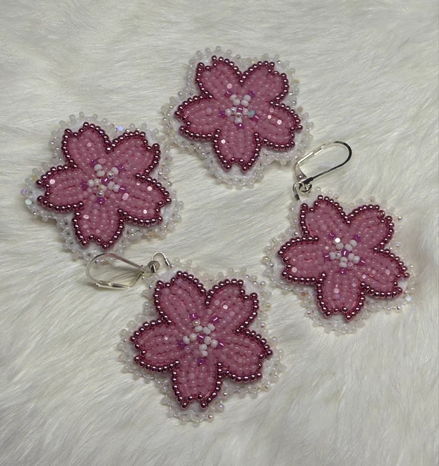 Flat stitched beaded earrings in a pink cherry blossom design. One pair is on posts and the other has lever hooks. Laying on white rabbit fur.