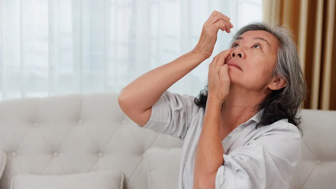 Middle-aged woman sitting on a beige couch applying eye drops to her right eye in a softly lit living room.