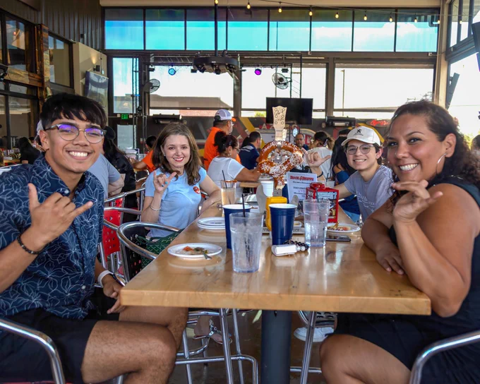 Smiling group of four people holding up the "Birds Up" hand gesture while sitting at a restaurant table with food and drinks. 