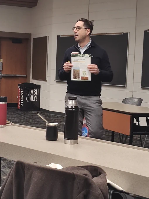 A man stands at the front of a room for a presentation. He is holding a factsheet handout.