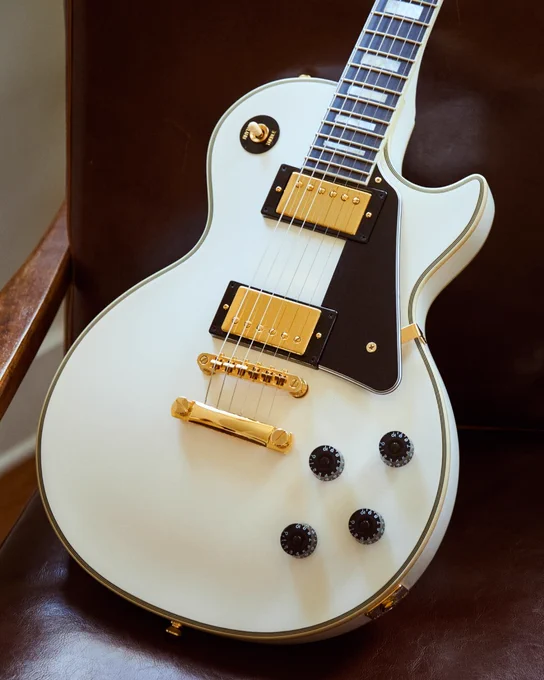 A white electric guitar, Epiphone Les Paul, is resting on a dark leather chair. The guitar features gold hardware, a black pickguard, and control knobs.