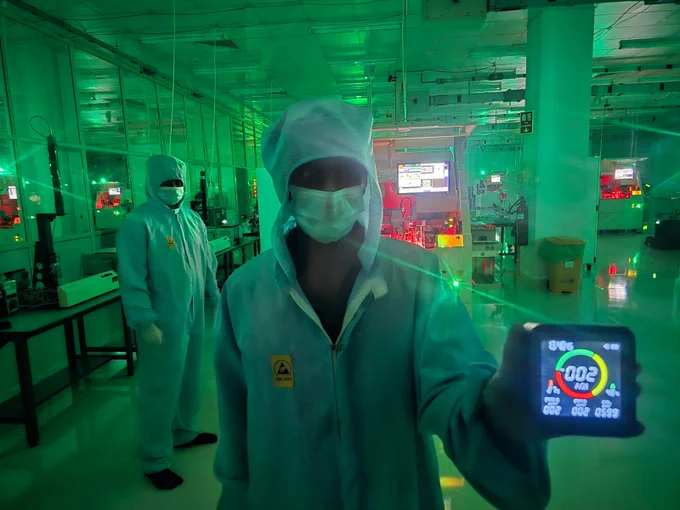 Two cleanroom technicians in full protective gear inside a semiconductor manufacturing lab, with green lighting and precision equipment. One displays an air quality monitor showing near-zero particulate levels.