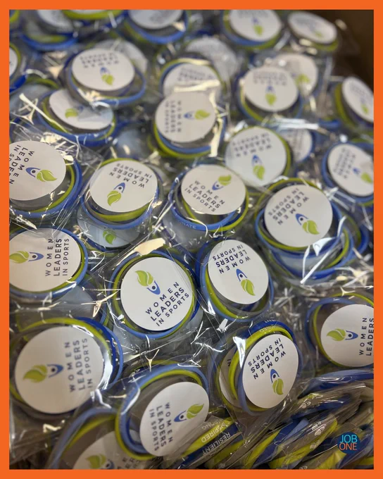 orange frame with a photo of green and blue rubber braceletes in clear packaging with Women Leaders in Sports logo stickers on each package. 