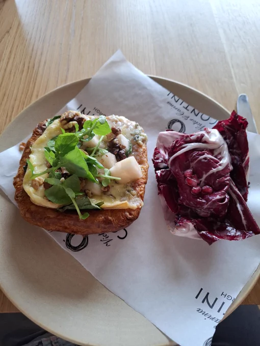 Grey plate with greaseproof paper. Sitting on this is an au gourmand buttery with blue cheese, pickled pears, toasted walnuts with a radicchio salad and pomegranite