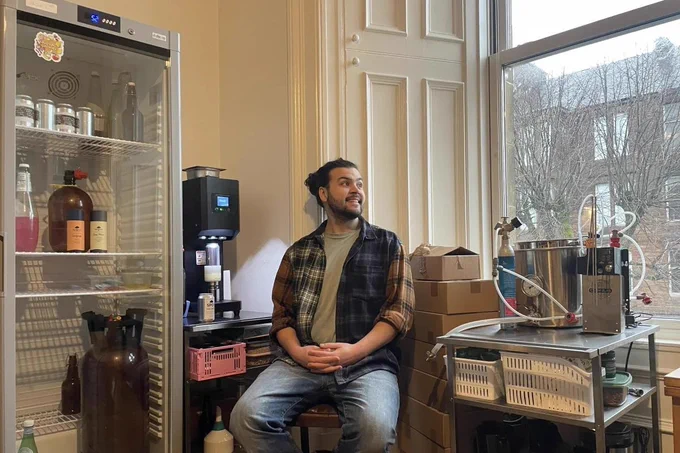Kane Edwards sitting on a stool in a small room with brewing equipment and a refrigerator filled with bottles and jars.