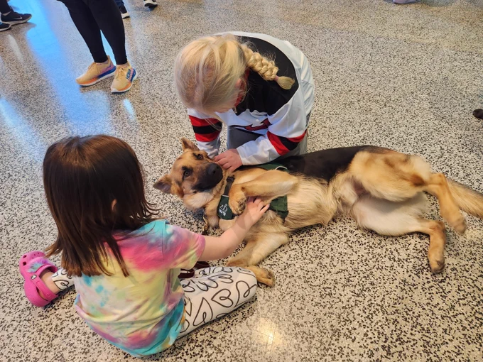 A German shepherd wearing a Seeing Eye puppy raising program vest lies on the floor of the Prudential Center lobby and gets belly rubs from two young girls who sit on the floor beside him.