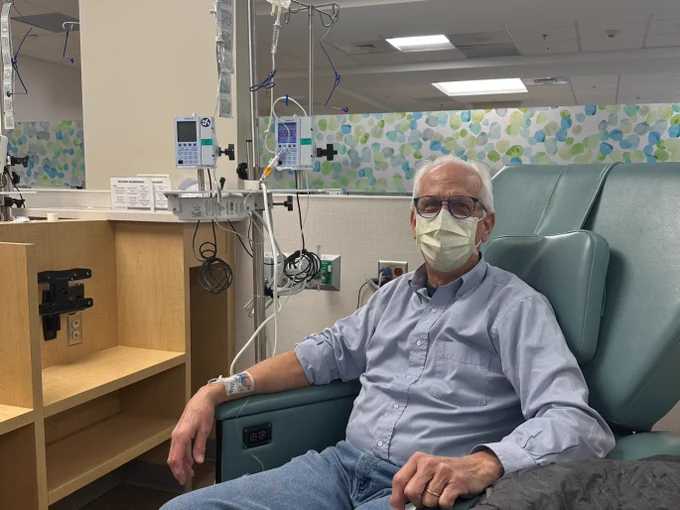An older man wearing a face mask is seated in a recliner-style chair in an outpatient hospital setting, receiving an IV treatment. Medical equipment and monitors are visible nearby. The man is wearing glasses and dressed in a light blue shirt and jeans, with a jacket draped over the chair's arm.