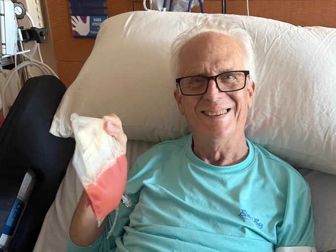 An older man with glasses is sitting up in a hospital bed, smiling and holding a stem cell transplant bag. Various medical devices are visible in the background, including monitors and a blood pressure cuff.
