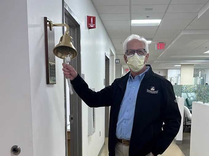 An older man wearing a face mask is standing in a hospital hallway, ringing a wall-mounted bell to signal the end of treatment. He is casually dressed in a blue shirt, black jacket, and khaki pants. 