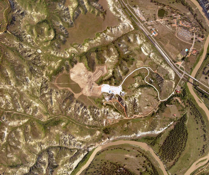 Aerial view of a winding river curving through rugged terrain with sparse vegetation and a construction site near a road.