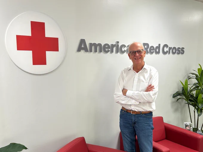 An older man stands smiling with his arms folded in a room with the American Red Cross logo on the wall. There are red chairs and green plants nearby.