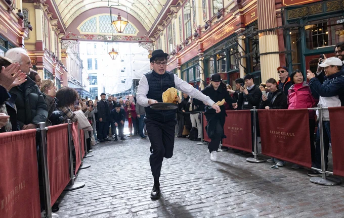 Young man in bowler hat races down a cobbled lane flipping a pancake in a pan whilst people either side watch on