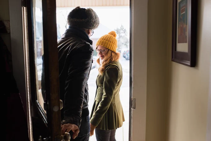 Two people standing in a doorway, one is wearing a yellow beanie. They are smiling and bathed in sunlight, creating a warm, welcoming atmosphere.