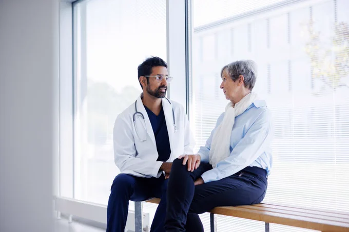 A medical professional in a white coat is having a consultation with a patient in a clinic beside a large window.
