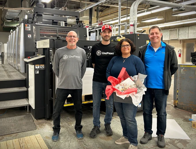 UniPrint customer service reps and press operators stand next to the offset printing press with a basket of #BuckeyeLove cookies from CHRR at The Ohio State University. 