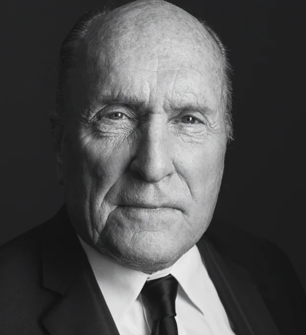 A close up head shot of Robert Duvall. Duvall is wearing black tie and looking thoughtfully towards the camera. The image is in black and white. 