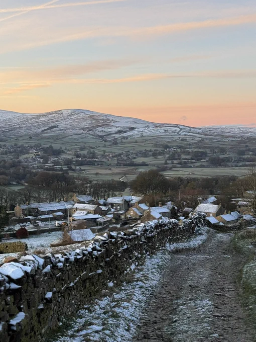 A winter rural landscape at sunrise, showing a snow-dusted stone lane leading downhill toward a small village of stone houses. The rooftops and surrounding dry‑stone walls are lightly covered in snow. Beyond the village, wide rolling hills stretch into the distance under a soft pink and orange sky.