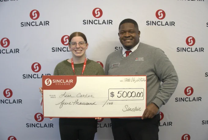Two people holding a large check for $5000 from Sinclair College. The background features a wall with repeating Sinclair College logos.