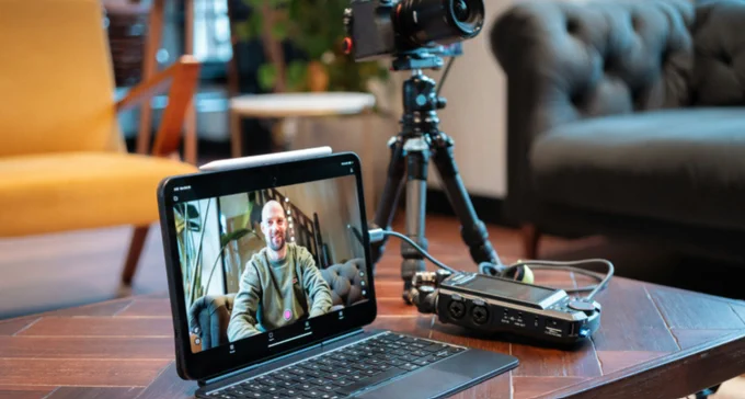 Video recording in progress on a laptop showing a person sitting on a couch, with a camera and audio equipment set up nearby on a wooden table.