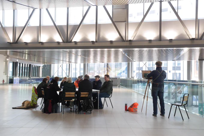 An artist sketches at an easel while a group sits together at a table in a large, open station concourse.