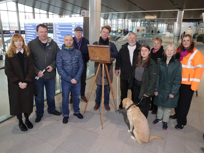 A group stands together inside a station concourse around an artist\u2019s easel, with a guide dog sitting at the front.
