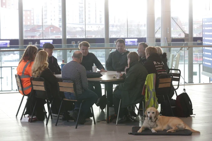 A group of people sit around a table in a bright, open station concourse, with a guide dog resting on a mat nearby.