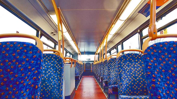 Interior view of an empty city bus with blue patterned seats and orange handrails under bright lighting.