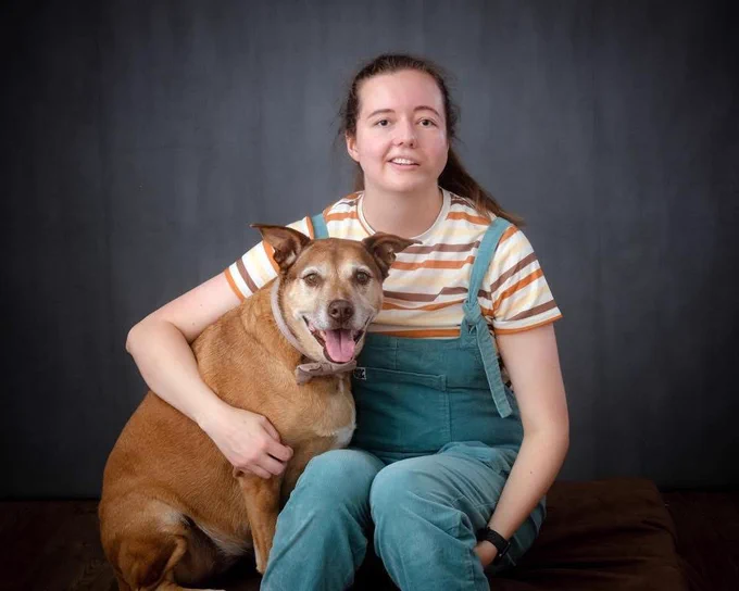 Chloe is a white woman in her twenties. it's a professional shoot of her and her dog, a large golden labrador. Chloe wears a striped t-shirt and pale blue Lucy and Yak dungarees.