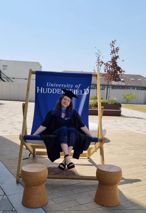 chloe sat on a large deck chair with university of huddersfield printed on it.  She's in her graduation outfit