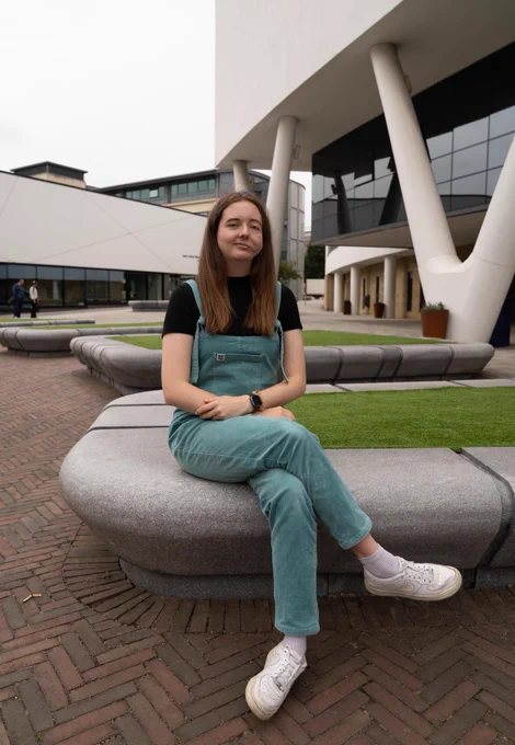 Chloe sat outside a large white and glass-fronted building in the same dungarees, smiling at the camera.