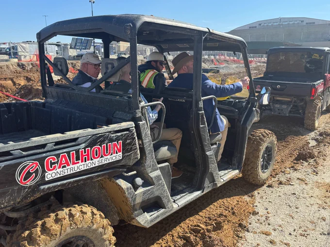 Commissioner Holt rides in a side-by-side utility vehicle at a muddy construction site with several men wearing safety gear.