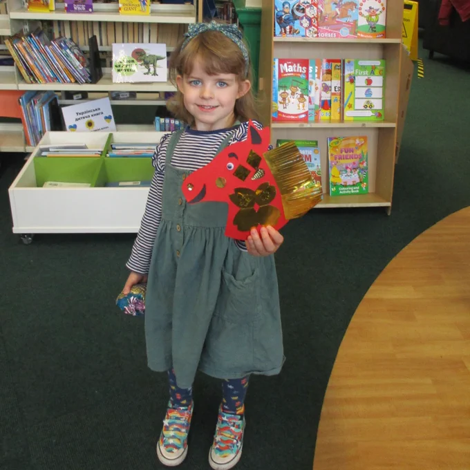 A child is standing in the library holding up the red and gold paper horse she made for Lunar New Year at story time