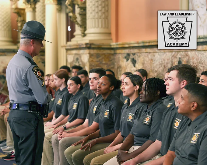 Cadets wearing polos and khakis are seated in the rear of the PA State House of Representatives chamber while a Trooper talks to them, plus the Law and Leadership program logo