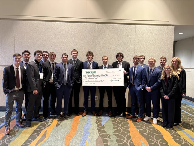 Purdue Turf students at GCSSA standing together indoors holding a large ceremonial check for the 2026 Collegiate Turf Bowl award.