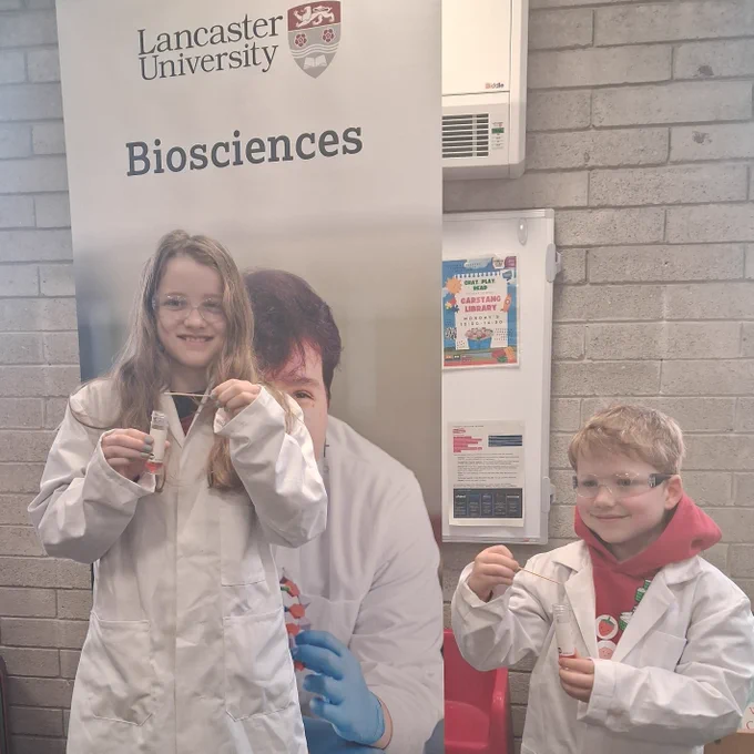 Two smiling children dressed in white lab coats and goggles, showing a science experiment in front of a Lancaster University banner