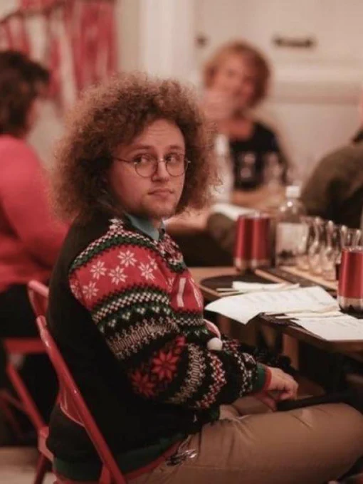 A man with curly brown hair, glasses and wearing a winter-themed sweater, sits in a chair at a table, looking over his right shoulder.
