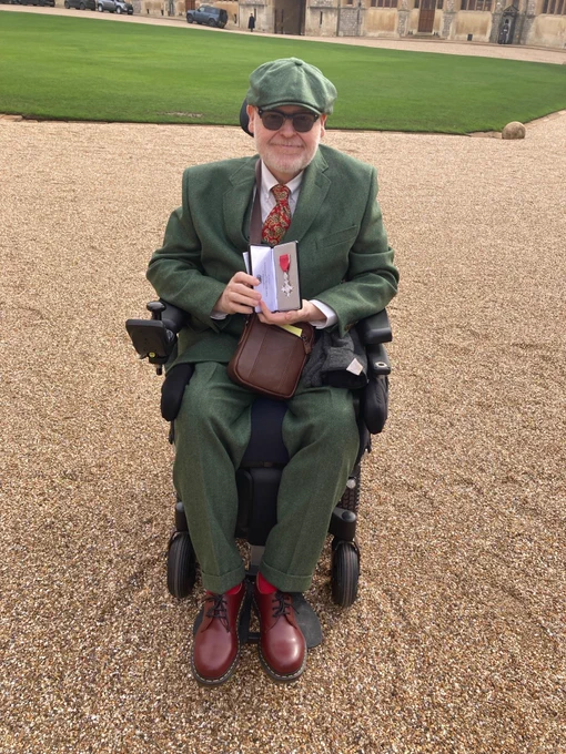 a man in a green suit and hat, in a wheelchair, holds an MBE medal in a box.