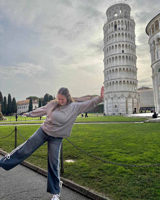 A person playfully poses with one leg raised, appearing to hold up the Leaning Tower of Pisa in the background. The scene is set in a grassy area with a paved path and other historic buildings nearby.